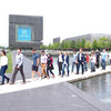 A large group of young adults walk across a footbridge over an expanse of water in front of the thyssenkrupp headquarters building