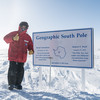 A man in a thick red jacket standing next to a plaque in the snow that reads "Geographic South Pole"