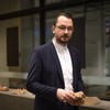 A man in a suit stands in front of a glass front and holds a wooden brick in his hands