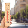 Eine Hand hält ein Thermometer, das etwa 30 Grad Celsius anzeigt. Im Hintergrund verschwommen eine Stadt im Sommer.