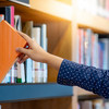 Ein Student zieht mit der rechten Hand ein oranges Buch aus einem Holzregal heraus