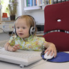 A toddler wearing headphones is sitting in front of a computer.